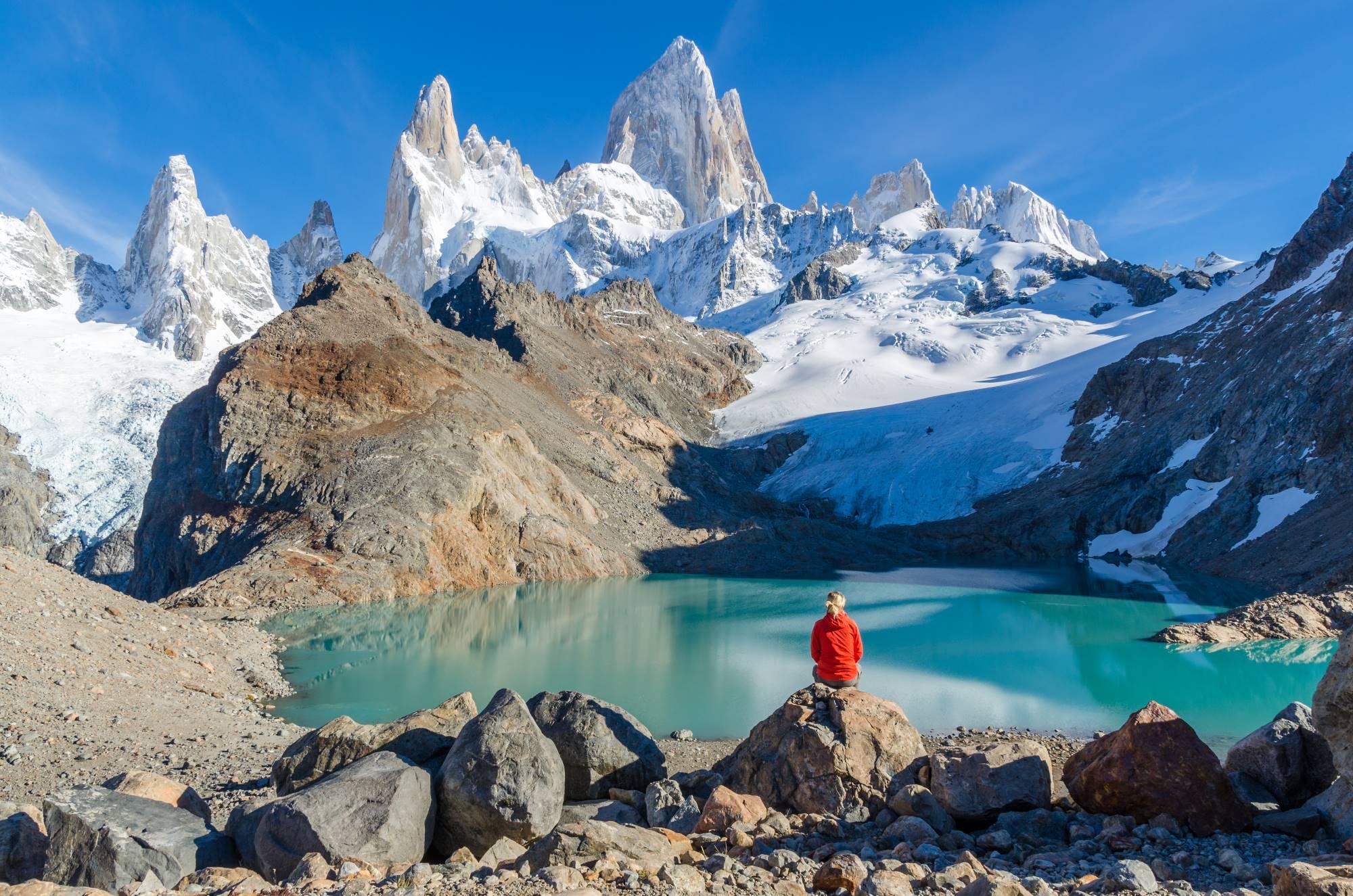 Turistka sedi na skali nad jezerom v naravnem parku Los Glaciares v Argentini s pogledom na špičasto goro FitzRoy.