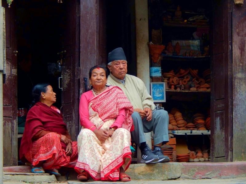 Starejši Nepalec in dve Nepalki v tradicionalnih nepalskih oblačilih posedajo pred eno od trgovin s keramičnimi izdelki.
