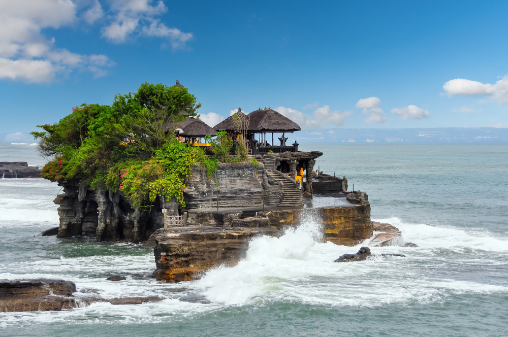 Pogled z obale na tempelj Tanah Lot in valove, ki udarjajo ob skalnato steno otoka.
