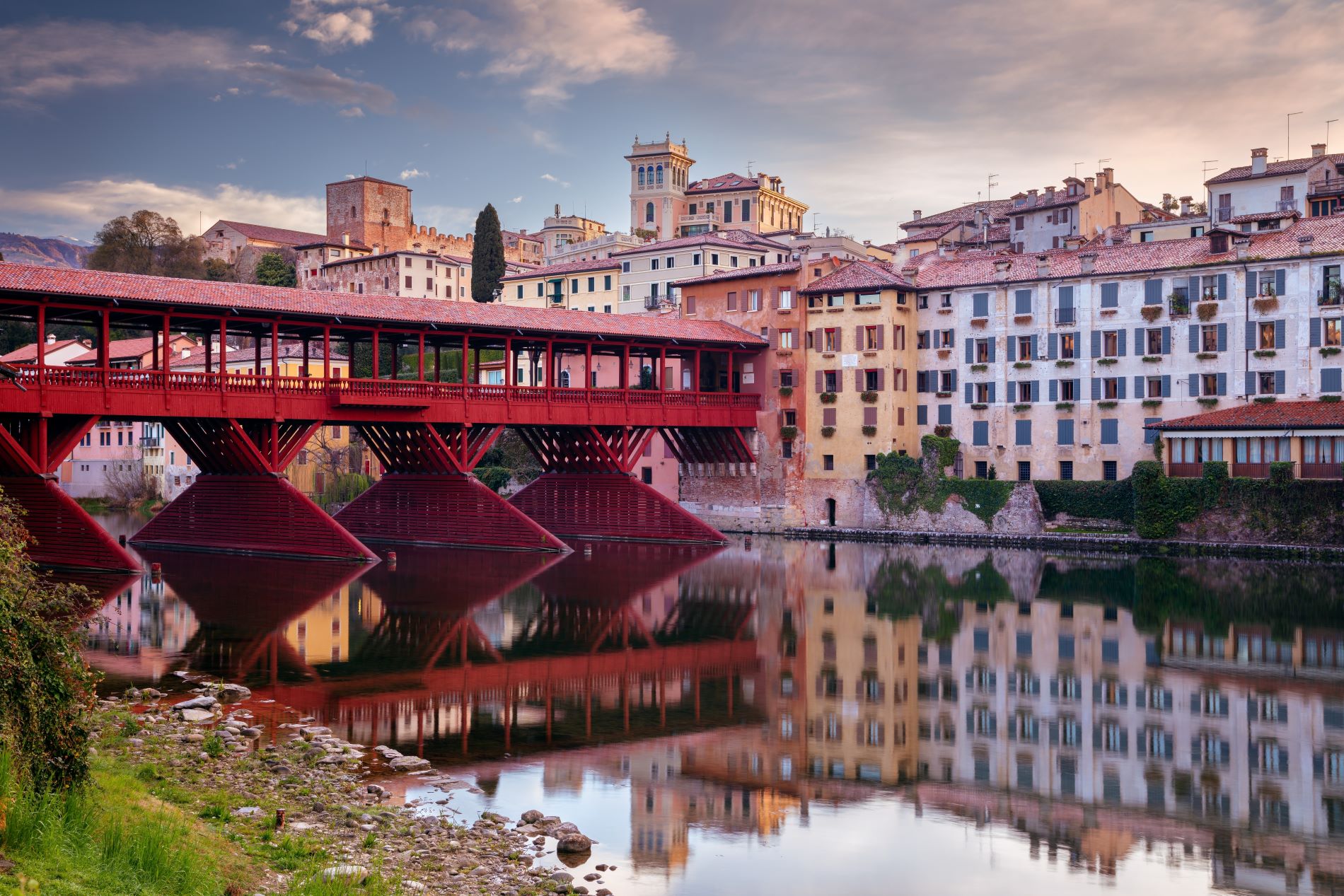 Pogled na leseni most Ponte Vecchio ali Ponte degli Alpini čez reko Brento v kraju Bassano del Grappa v severni Italiji v večerni svetlobi in pisane hiše starega mestnega jedra ob rečnem nabrežju.