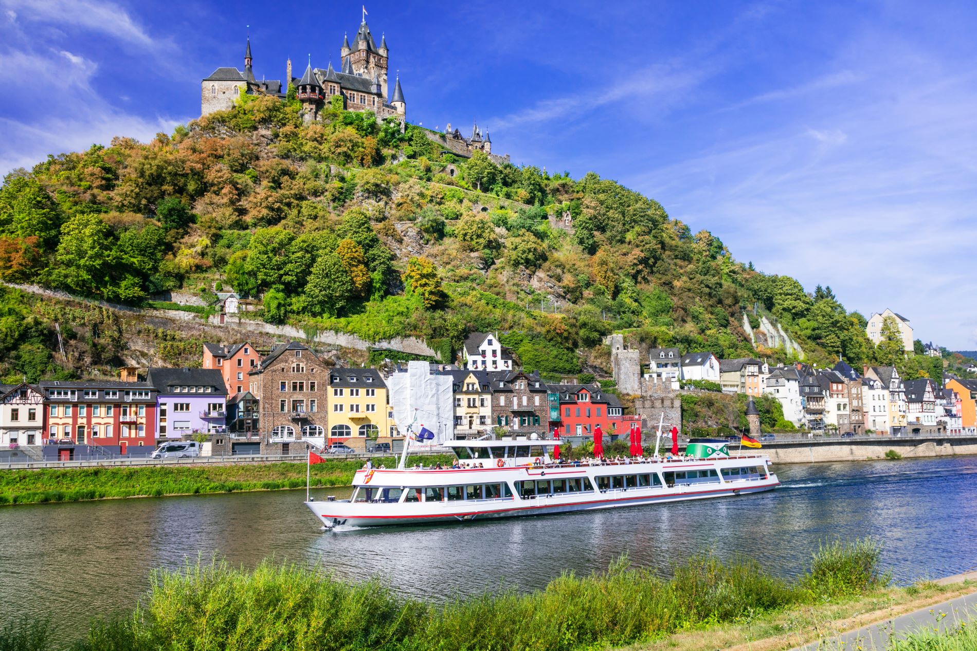 Pogled na mestece Cochem v Nemčiji z reke Mozele, po kateri pluje turistična ladja. Nad mestom z barvitimi hišami se dviga mogočen grad.