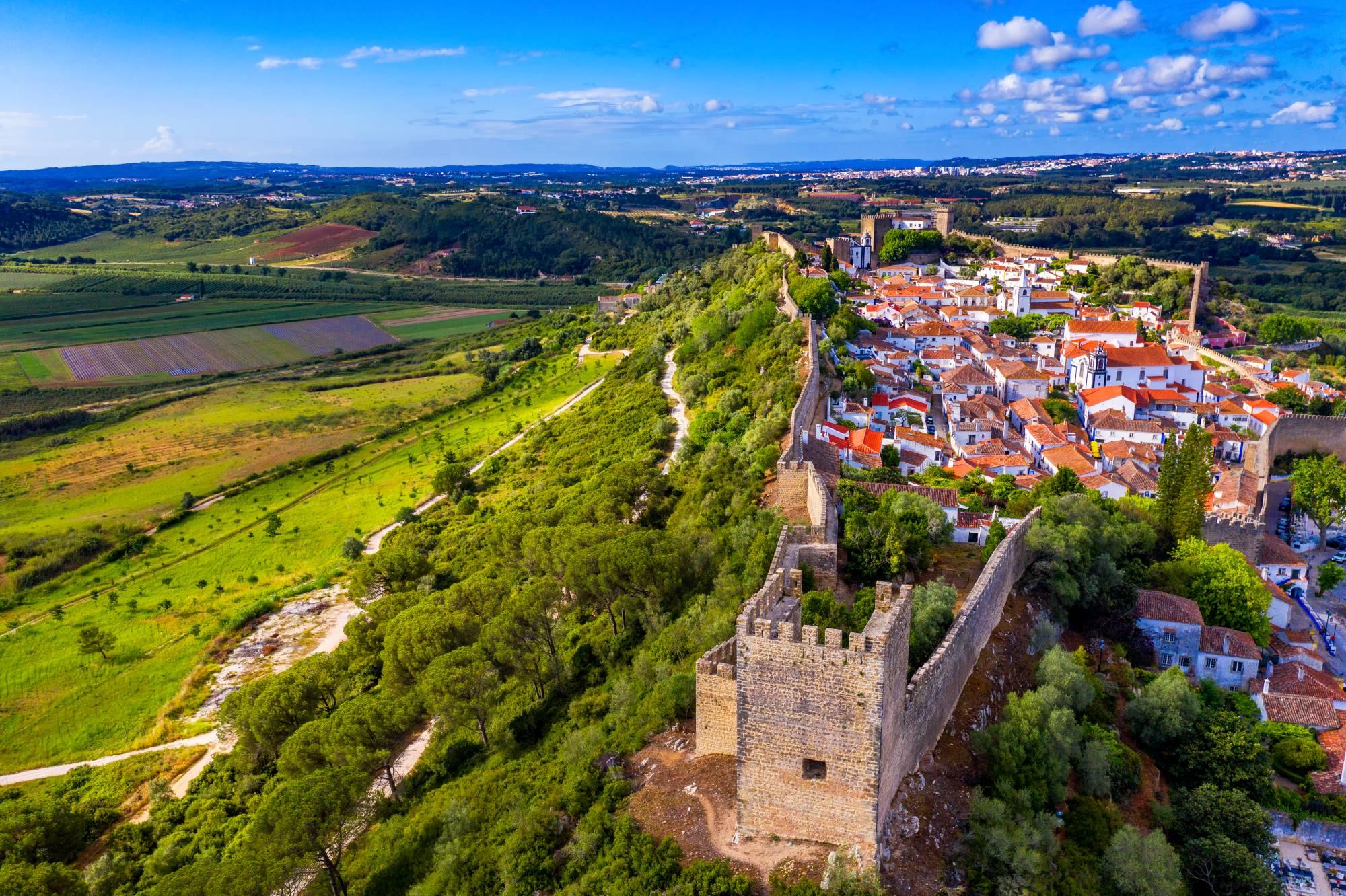 Panoramski pogled na z obzidjem obdano mestece Óbidos, ki se dviguje nad ozelenelo pokrajino Portugalske.