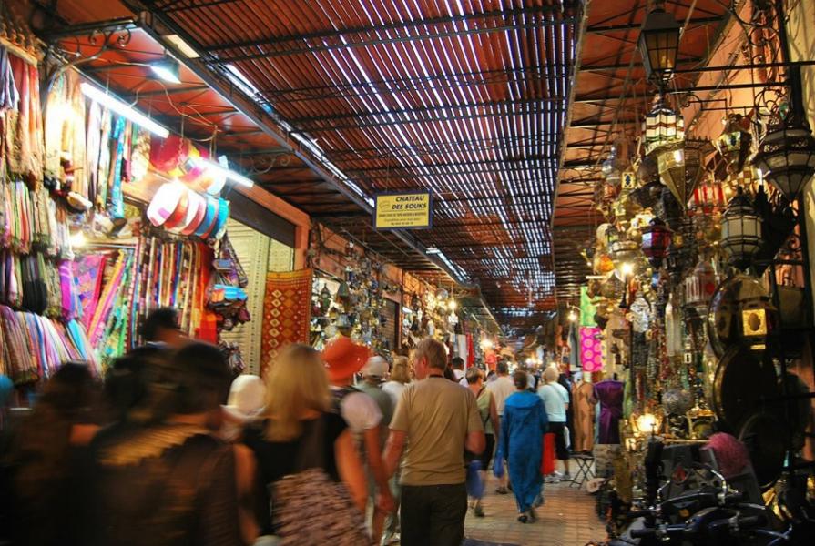 Turisti in domačini se sprehajajo skozi suk v Marakešu, kjer prodajalne ponujajo vse od blaga, oblačil in posode do luči in nakita.