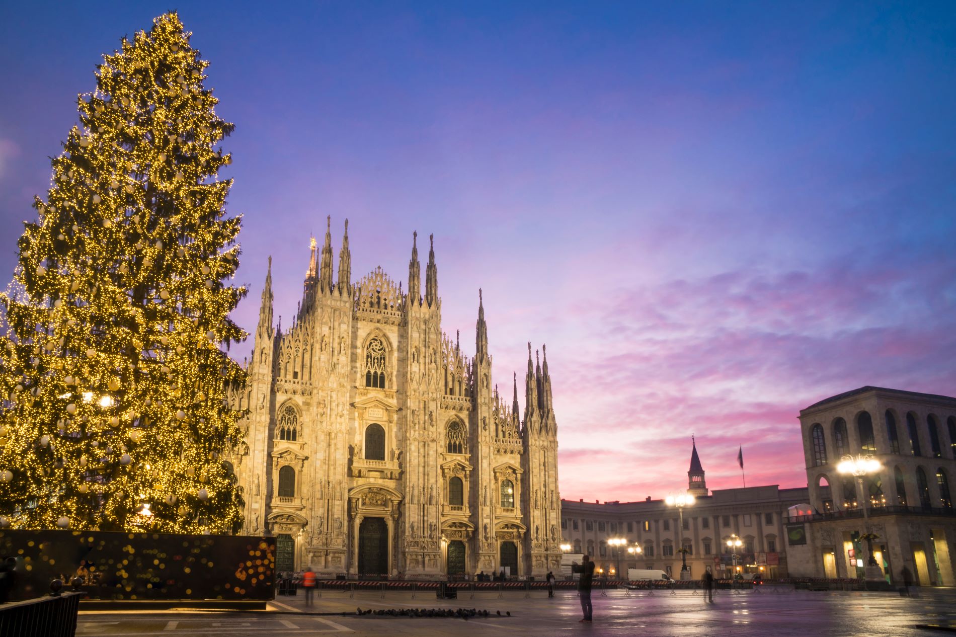Jutranja svetloba z rdečim žarom obdaja trg Piazza del Duomo v središču Milana, na katerem stoji mogočna gotska stolnica in osvetljeno božično drevo.