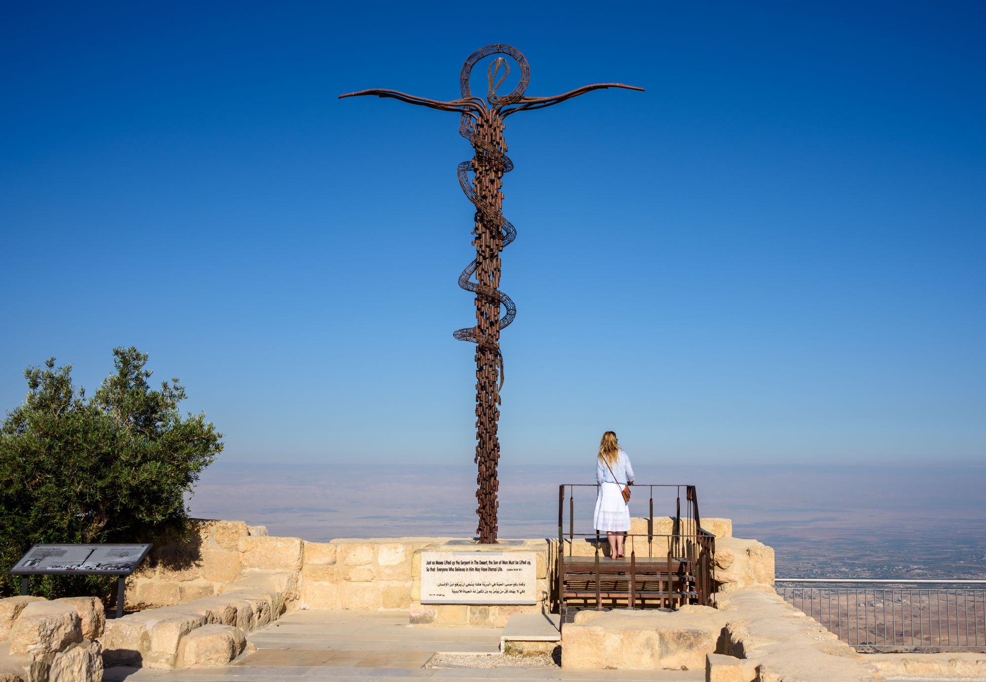 Obiskovalka na razgledni ploščadi pod kipom križa s kačo na gori Nebo v Jordaniji, od koder naj bi Mojzes prvič ugledal Obljubljeno deželo in od koder se odpira pogled na dolino reke Jordan, Mrtvo morje in celo do Jeruzalema.