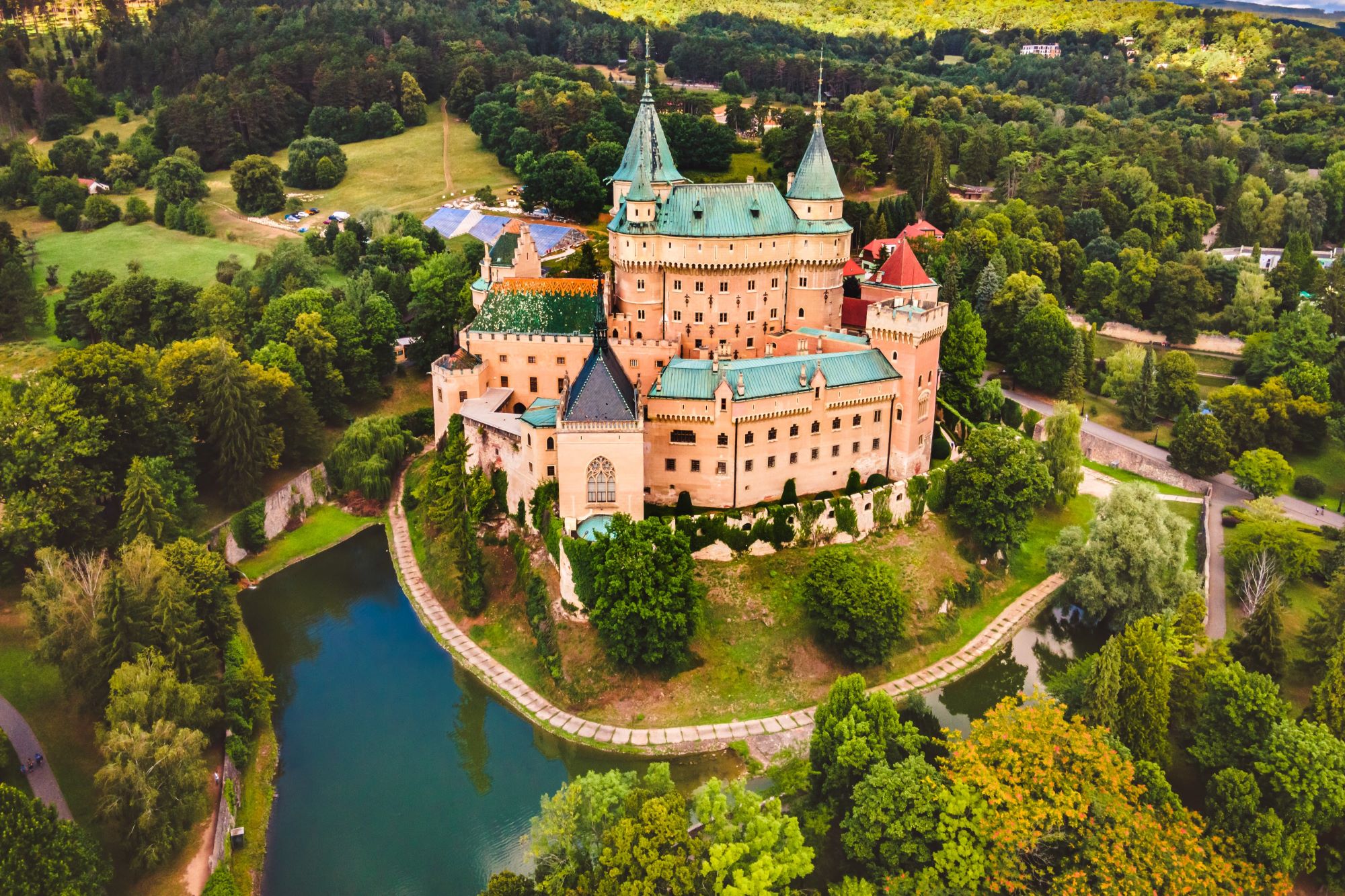 Pogled iz zraka na pravljično romantični grad v Bojnicah na Slovaškem z zunanjim in notranjim gradom ter tremi dvorišči, obdan z jezeron in zelenjem.