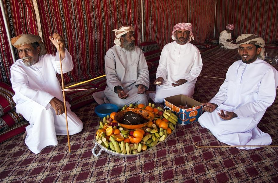 Omanski moški v tradicionalnih oblačilih, belih haljah dišdašah in s palestinsko ruto na glavi, v puščavi v šotoru pričakujejo turiste s polno mizo sadja.