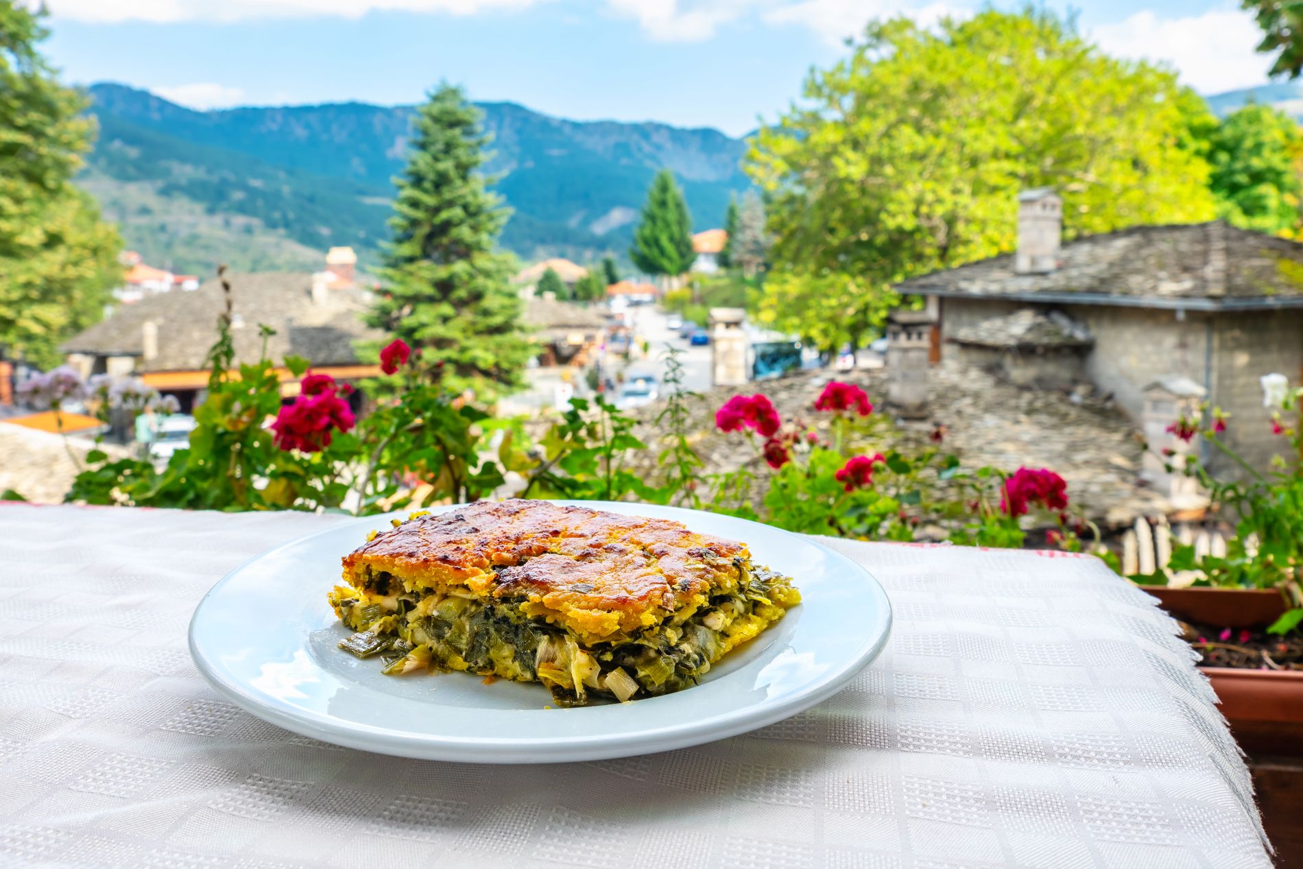 Krožnik z značilno grško pito iz listnatega testa s špinačo, imenovano spanakopita, in v ozadju panoramski pogled na slikovito vas na planoti Metsovo v severni Grčiji.